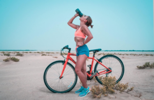 mujer de short azul y zapatos azules tomando agua entrenando en bicicleta